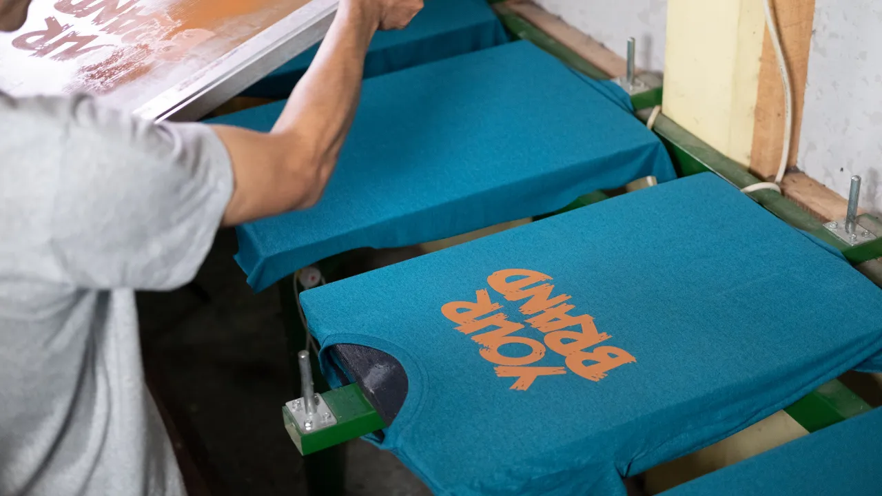 A worker using a screen for custom t shirt silk screen printing, pressing an orange brand logo onto a line of blue shirts.