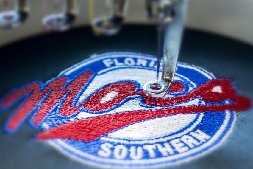 Embroidery vs. Screen Printing in action—machine stitching a colorful “Mocs” logo onto fabric, showcasing precision and texture unique to embroidery.